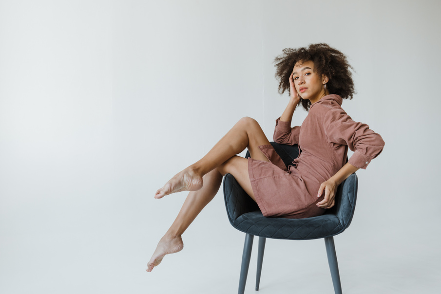 An Afro-Haired Woman in Brown Dress Posing while Sitting on the Chair